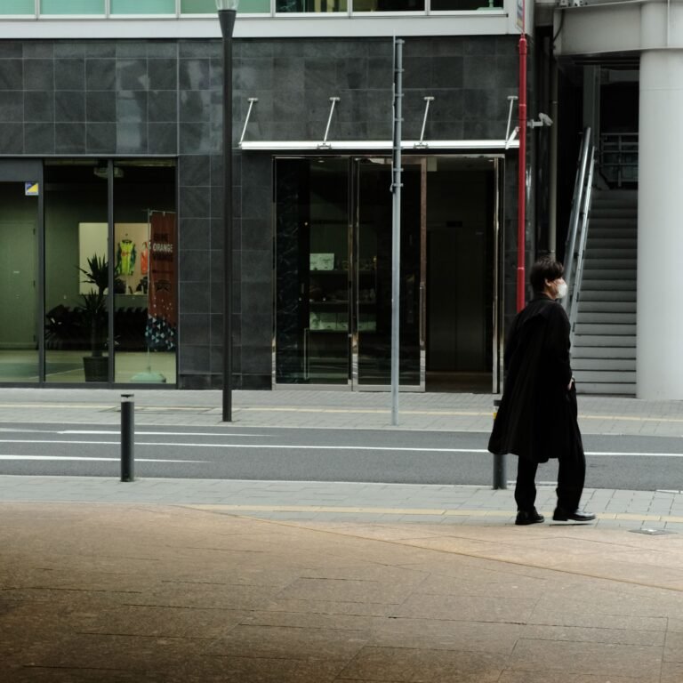 Home Man in dark coat and mask walking past modern building in urban setting.