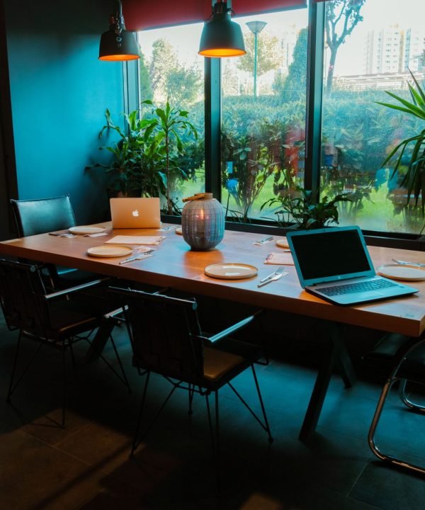 A modern dining room setup with laptops, plates, and lush plants by large windows.